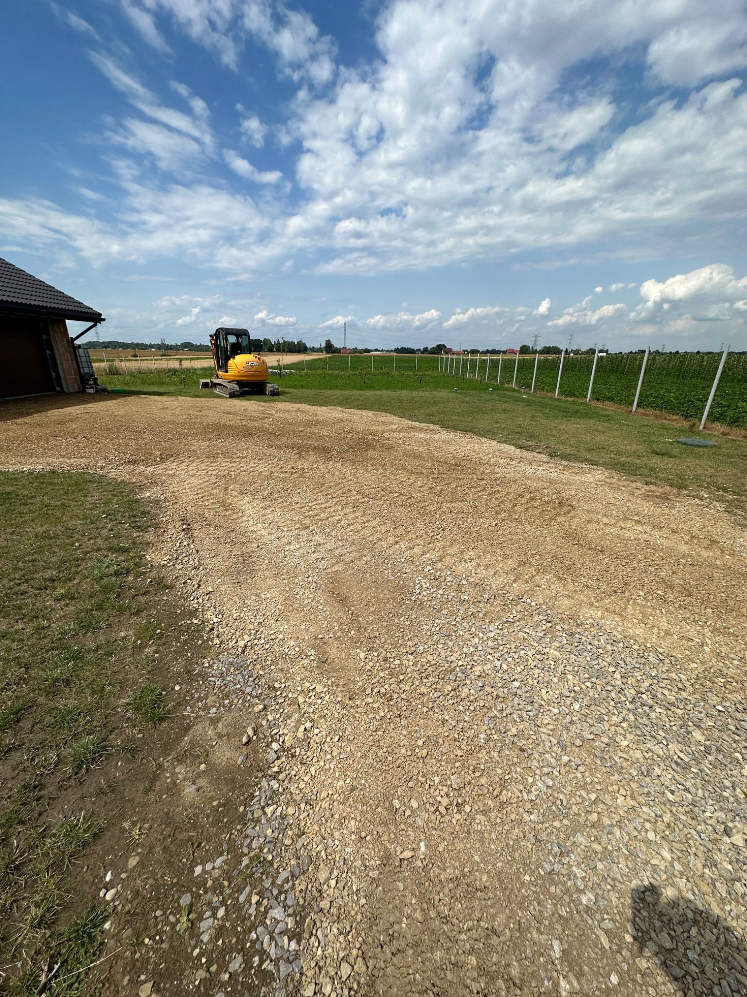 Żółta mini koparka JCB na utwardzonym żwirowym placu budowy, w tle budynek z garażem i ogrodzenie pola uprawnego pod błękitnym, obłocznym niebem.