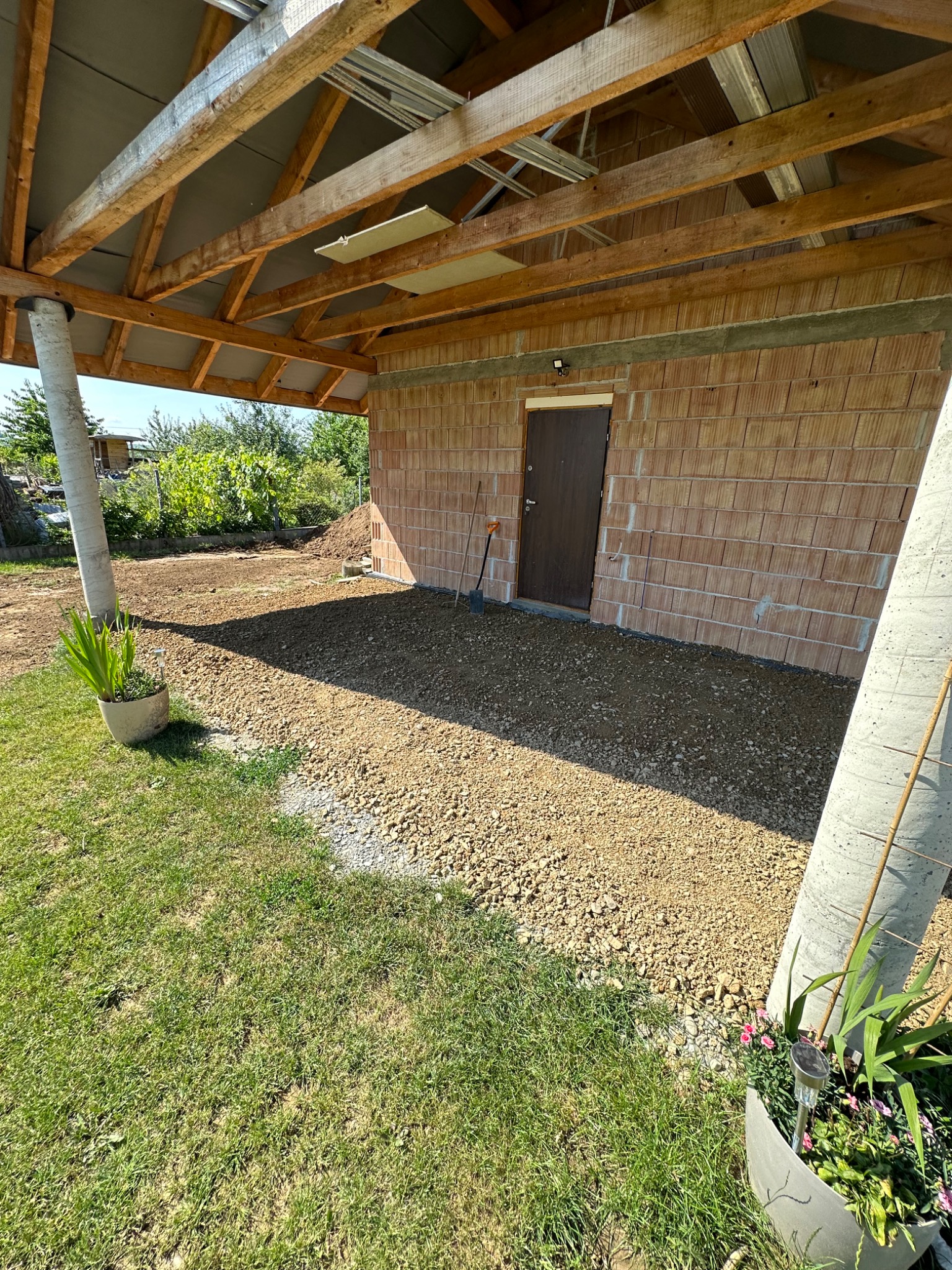 Przygotowany teren pod wiatą z ceglaną ścianą, wyrównany żwirem, z widocznymi słupami podtrzymującymi dach i roślinami w donicach na pierwszym planie.