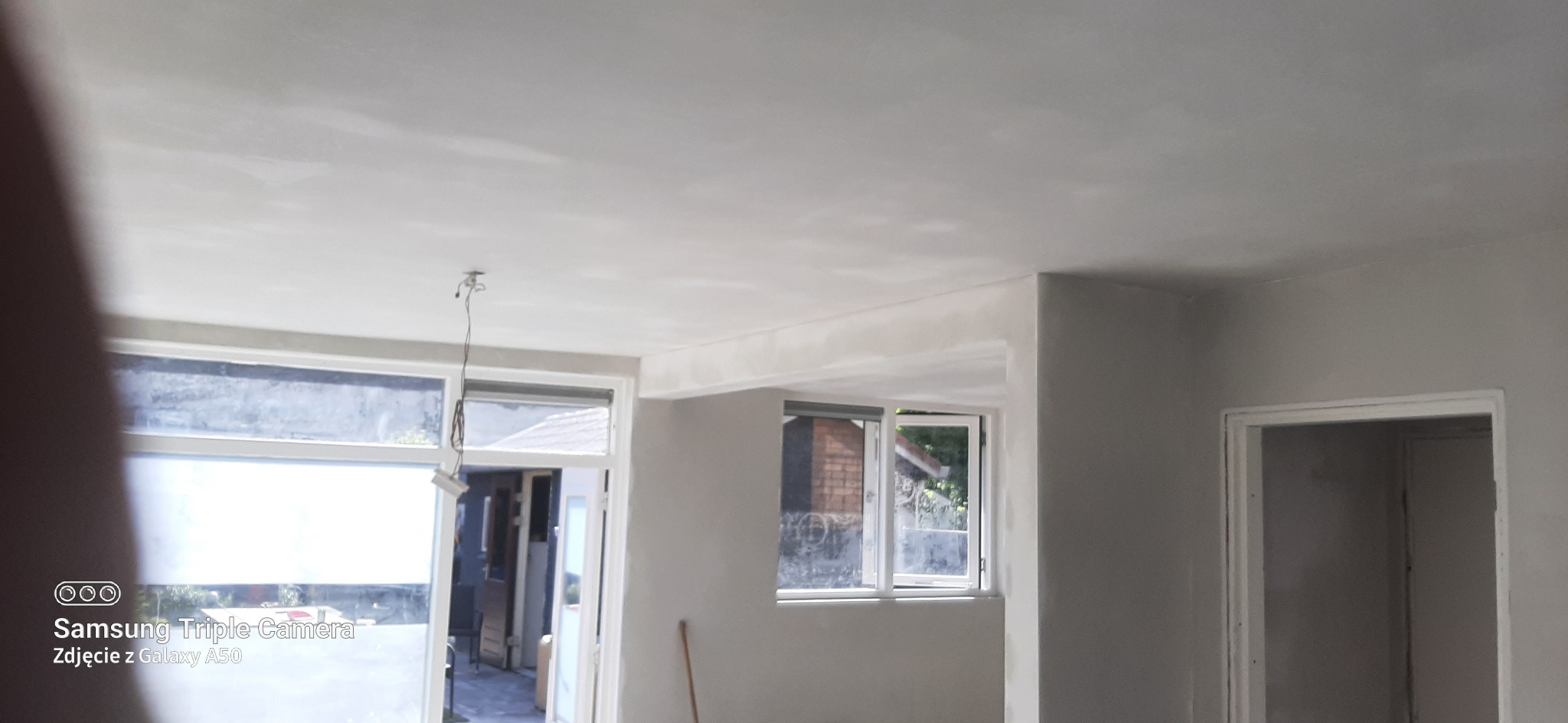 Wnętrze pokoju po nałożeniu gładzi szpachlowej na ścianach i suficie, widoczne okna z roletami i drzwi do innego pomieszczenia, surowy stan wykończenia.