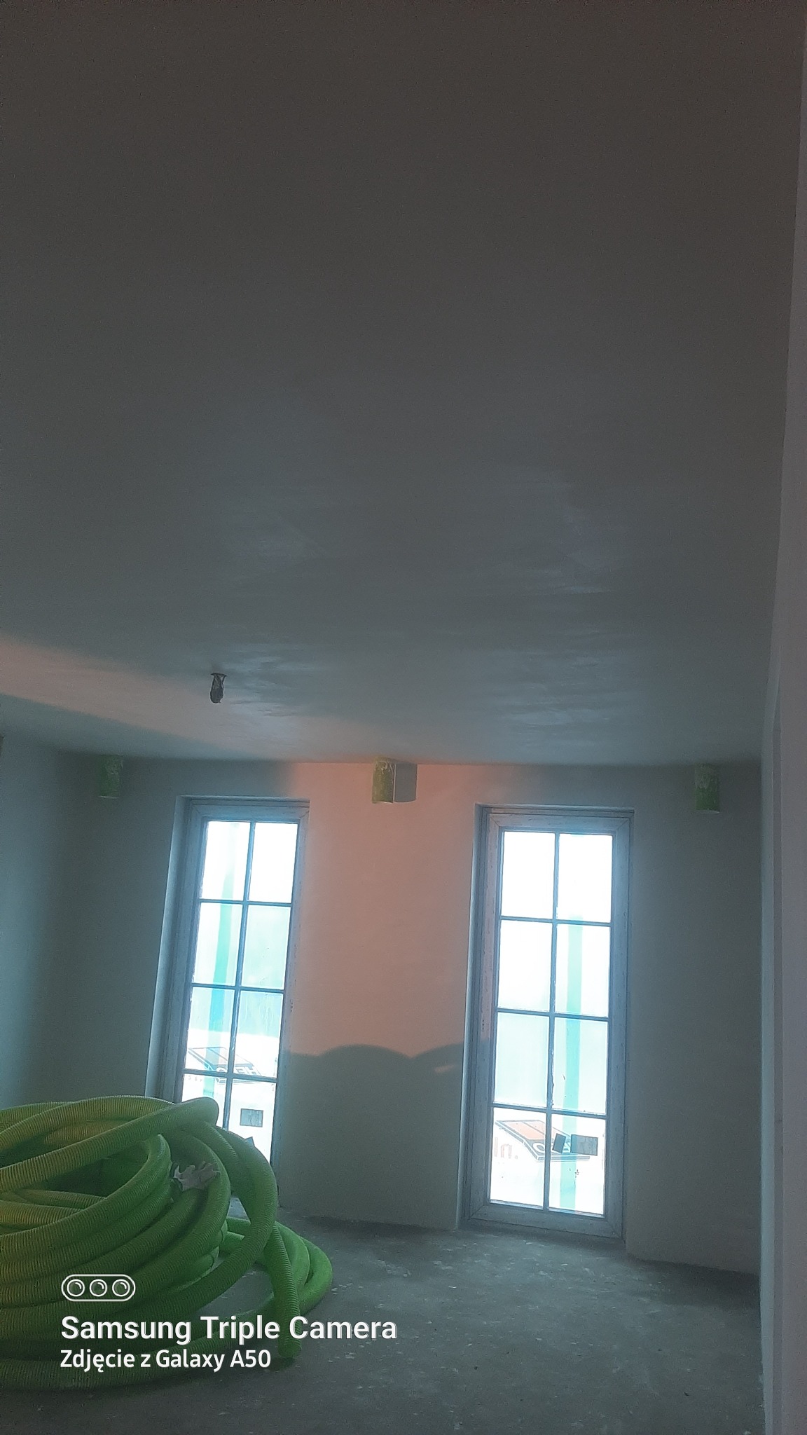 Wnętrze pokoju w trakcie prac wykończeniowych: ściany przygotowane pod malowanie, zabezpieczone okna, na podłodze zwój zielonego węża wentylacyjnego.