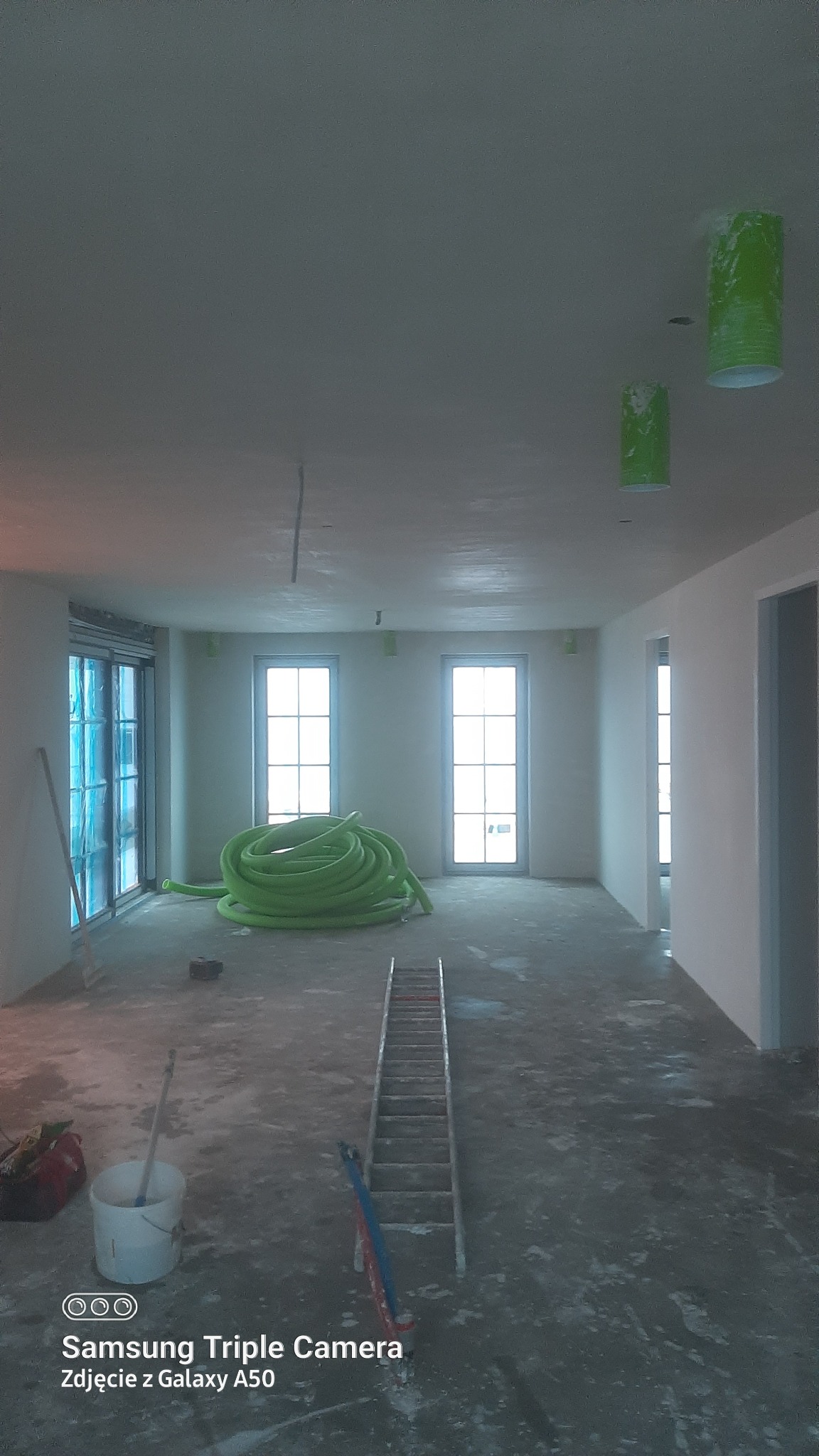 Wnętrze w trakcie prac przygotowawczych: zabezpieczone okna folią, zwój zielonego węża na podłodze, drabina, wiadro z narzędziami, sufit po szpachlowaniu, zabezpieczone lampy.