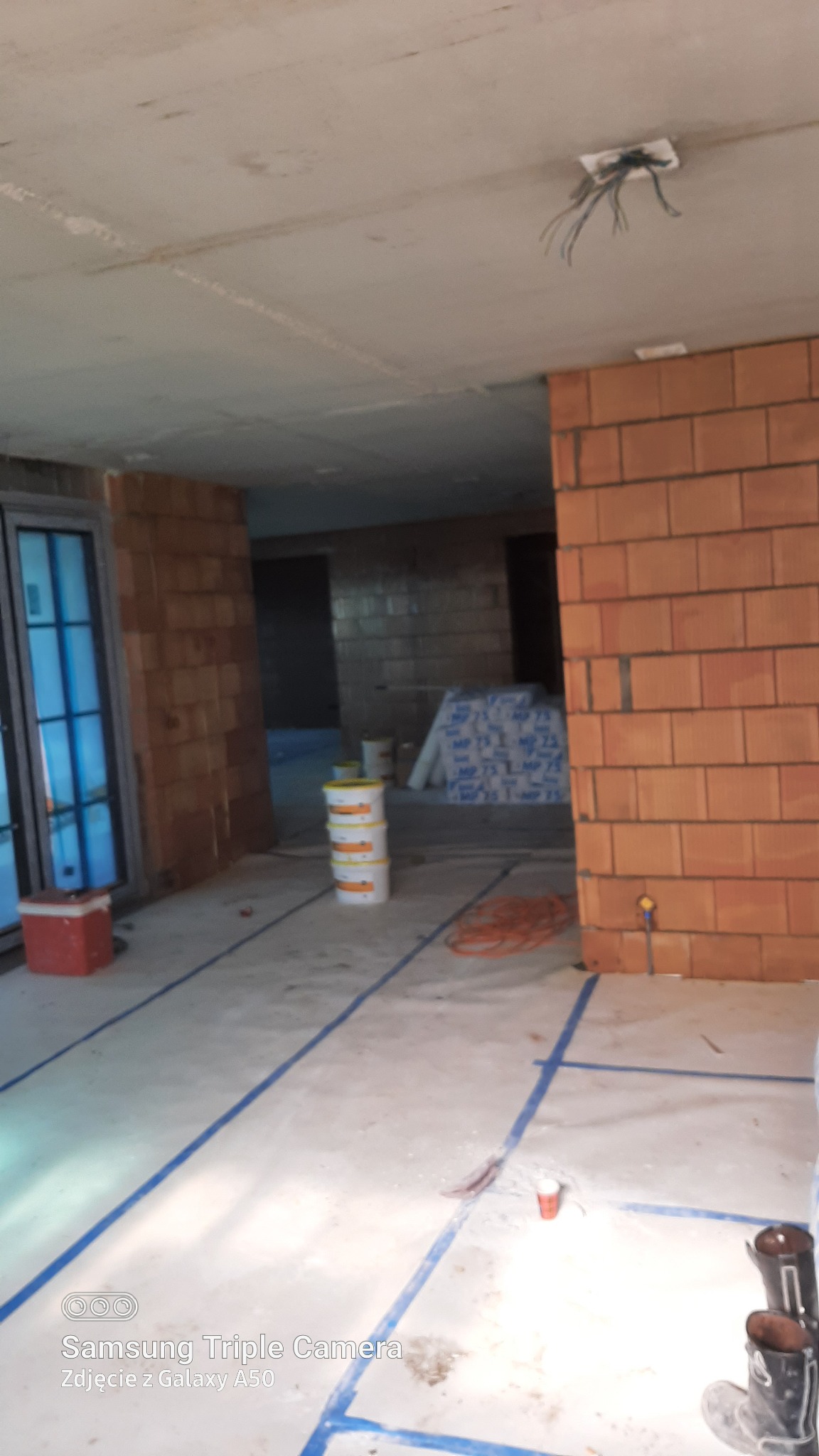 Wnętrze w surowym stanie przed wykończeniem, z widocznymi ścianami z cegły, przewodami elektrycznymi wystającymi z sufitu, zabezpieczoną folią podłogą wyklejoną niebieską taśmą, oraz ustawionymi...