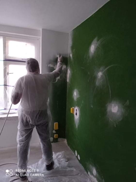 Pracownik w kombinezonie ochronnym natryskowo aplikuje białą farbę na zieloną ścianę z widocznymi miejscami po szpachlowaniu i gniazdkami elektrycznymi, podłoga zabezpieczona folią.