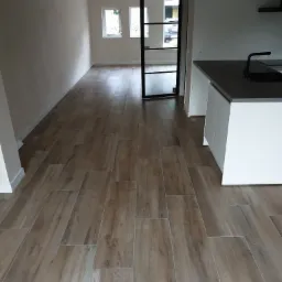 Ułożona podłoga z płytek imitujących drewno w jasnym wnętrzu, widoczny fragment zabudowy kuchennej i przeszklone drzwi do kolejnego pomieszczenia.