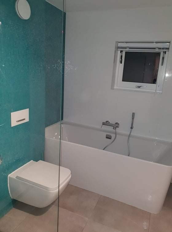 Nowoczesna łazienka z turkusową mozaiką, podwieszaną toaletą i białą wanną, widok przez szklaną ściankę kabiny.