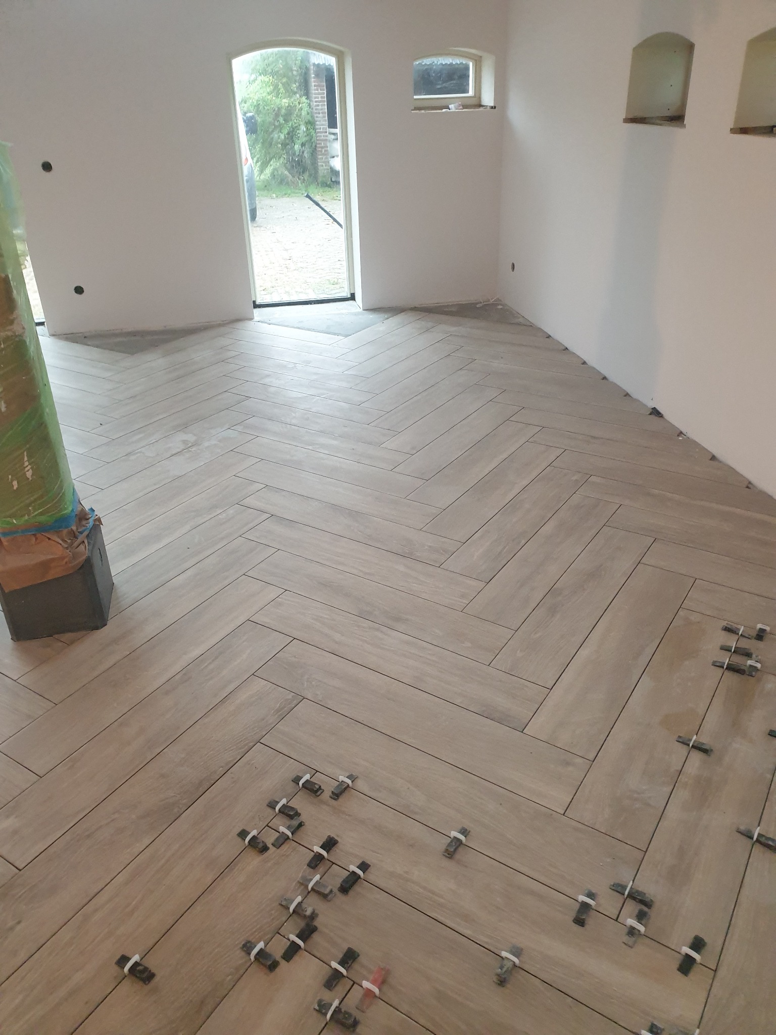 Układanie płytek podłogowych w jodełkę, widoczne plastikowe krzyżyki dystansowe, kolumna zabezpieczona folią malarską, świeżo wykończone ściany w jasnym pomieszczeniu z oknami.