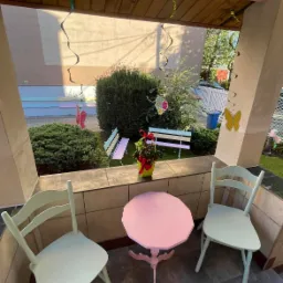 Pastelowy kącik zabaw na zewnątrz z różowym stolikiem i miętowymi krzesłami, dekoracje motylami i wstążkami, małe kolorowe ławeczki w tle.