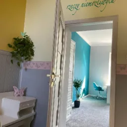 Wnętrze pokoju dziecięcego z otwartymi drzwiami, żółto-niebieskie ściany z motywami motyli, białe meble, roślina doniczkowa i napis 'Nie ma rzeczy niemożliwych'.