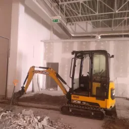 Żółta mini koparka JCB 19C-1 w trakcie prac ziemnych wewnątrz budynku w Wałbrzychu, widoczny wykop w ziemi i gruz.