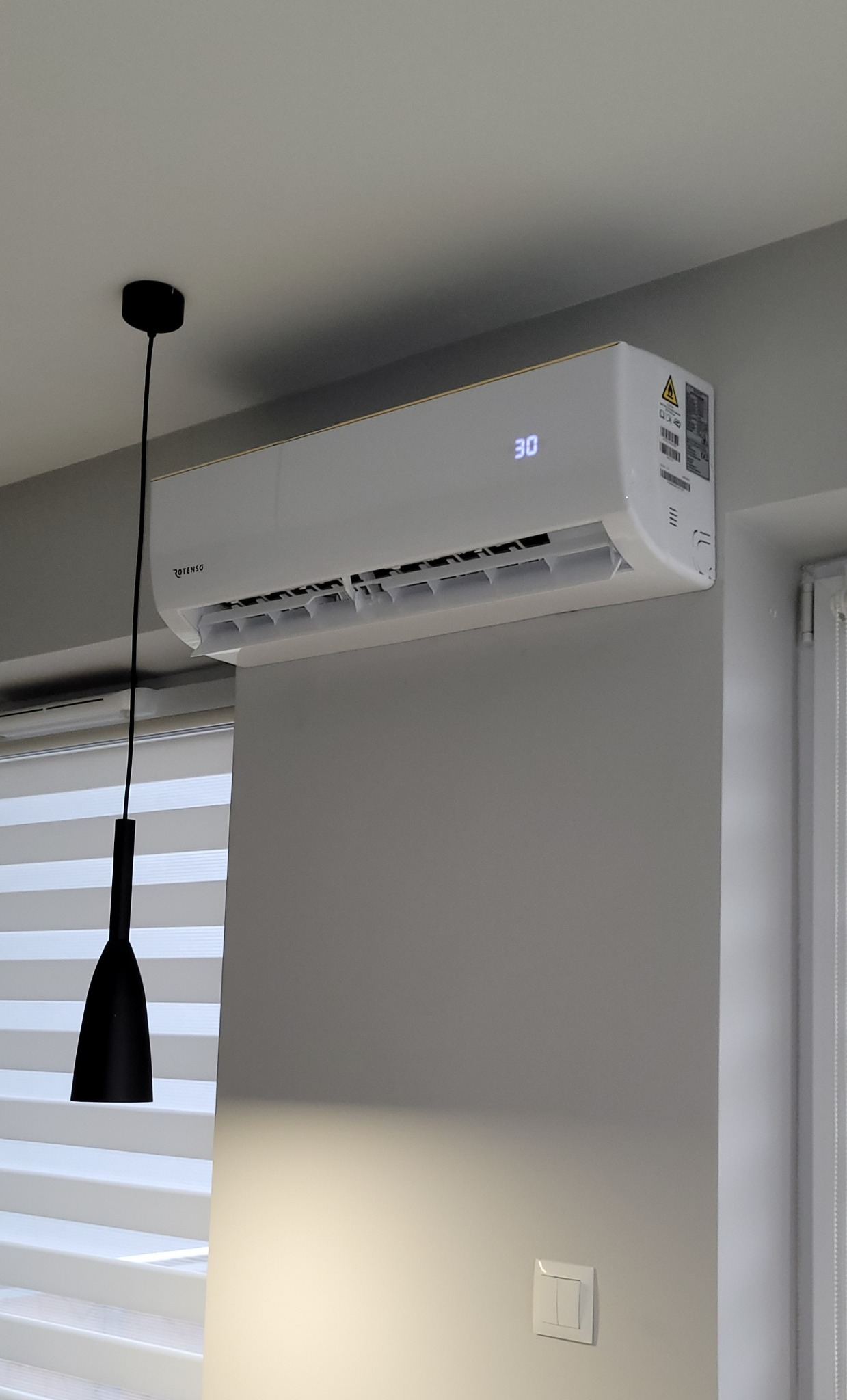 Biała klimatyzacja Rotenso na ścianie, z wyświetlaczem 30 stopni, obok okna z żaluzjami i czarną lampą wiszącą. Minimalistyczny design wnętrza.