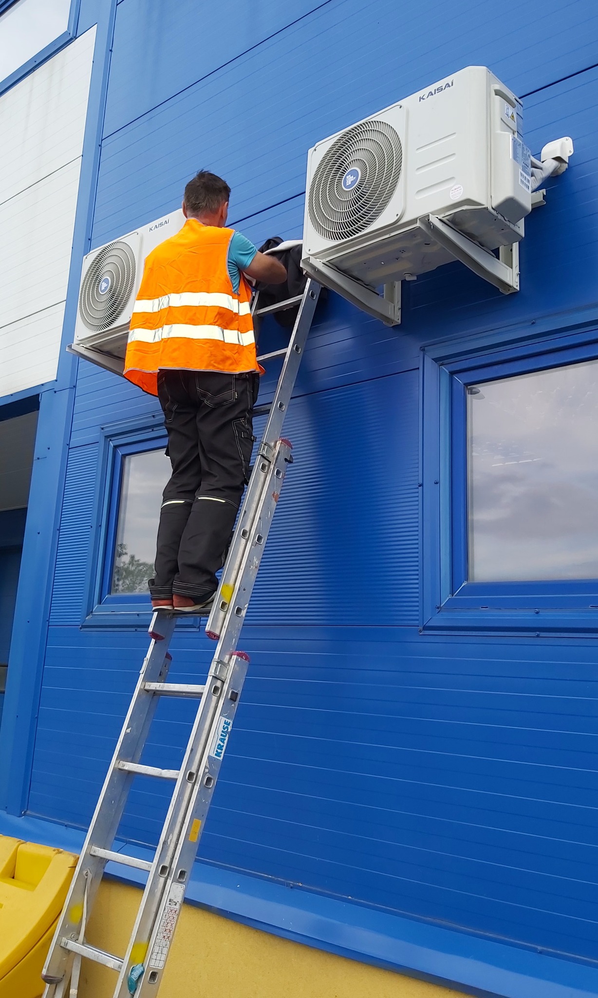 Montaż klimatyzacji Kaisai na niebieskiej fasadzie. Instalator w kamizelce na drabinie, widoczne dwa urządzenia zewnętrzne. Dwa okna w tle.