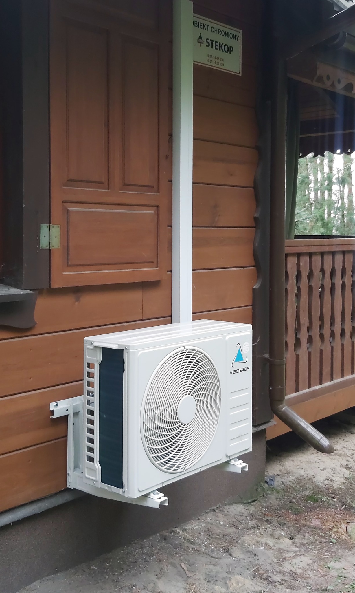 Klimatyzator Vesser zamontowany na drewnianej elewacji budynku z brązowymi okiennicami i rynną. Biała rura pionowa. Widoczny znak informacyjny 'Obiekt chroniony'.