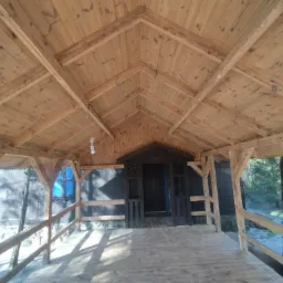 Drewniana altana z widoczną konstrukcją dachu, barierkami i wejściem, otoczona zielenią.