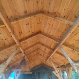 Drewniany sufit z widocznymi belkami konstrukcyjnymi, oświetlony wiszącą lampą w stylu rustykalnym, z fragmentem drzwi w tle.