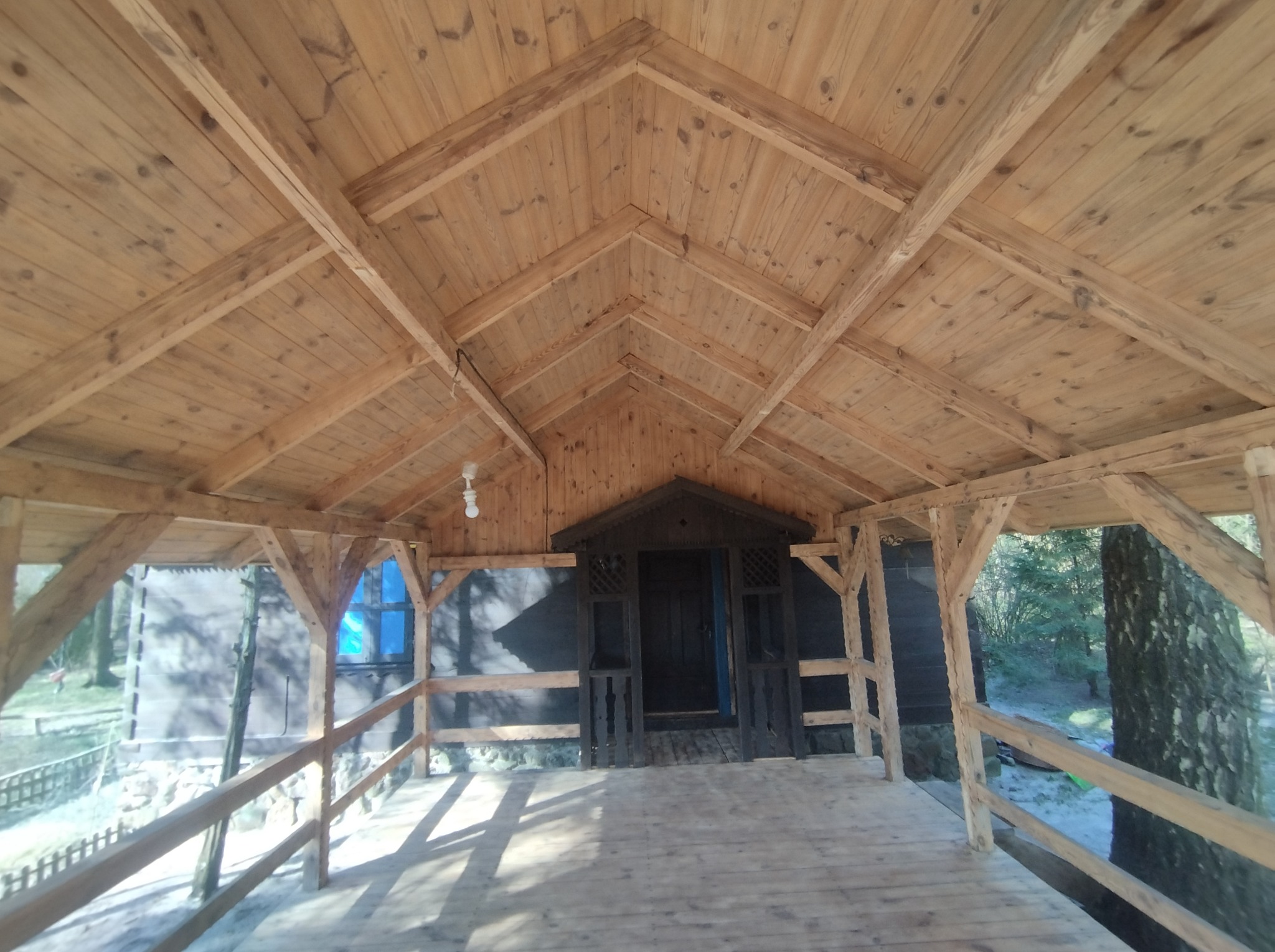 Drewniana altana z widoczną konstrukcją dachu, barierkami i wejściem, otoczona zielenią.