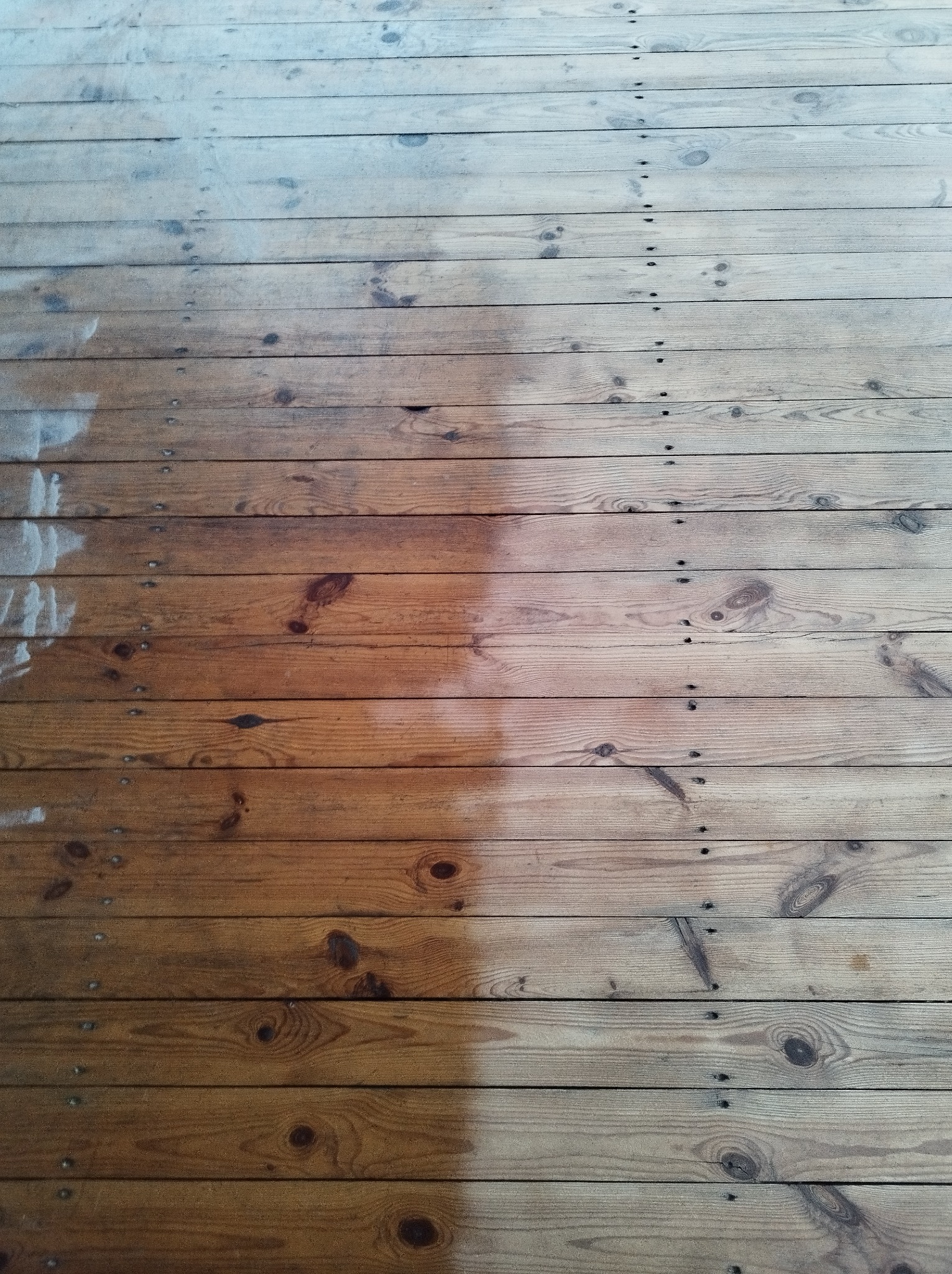 Drewniana podłoga z desek, częściowo wyczyszczona, ukazująca różnicę w kolorze i strukturze przed i po czyszczeniu.