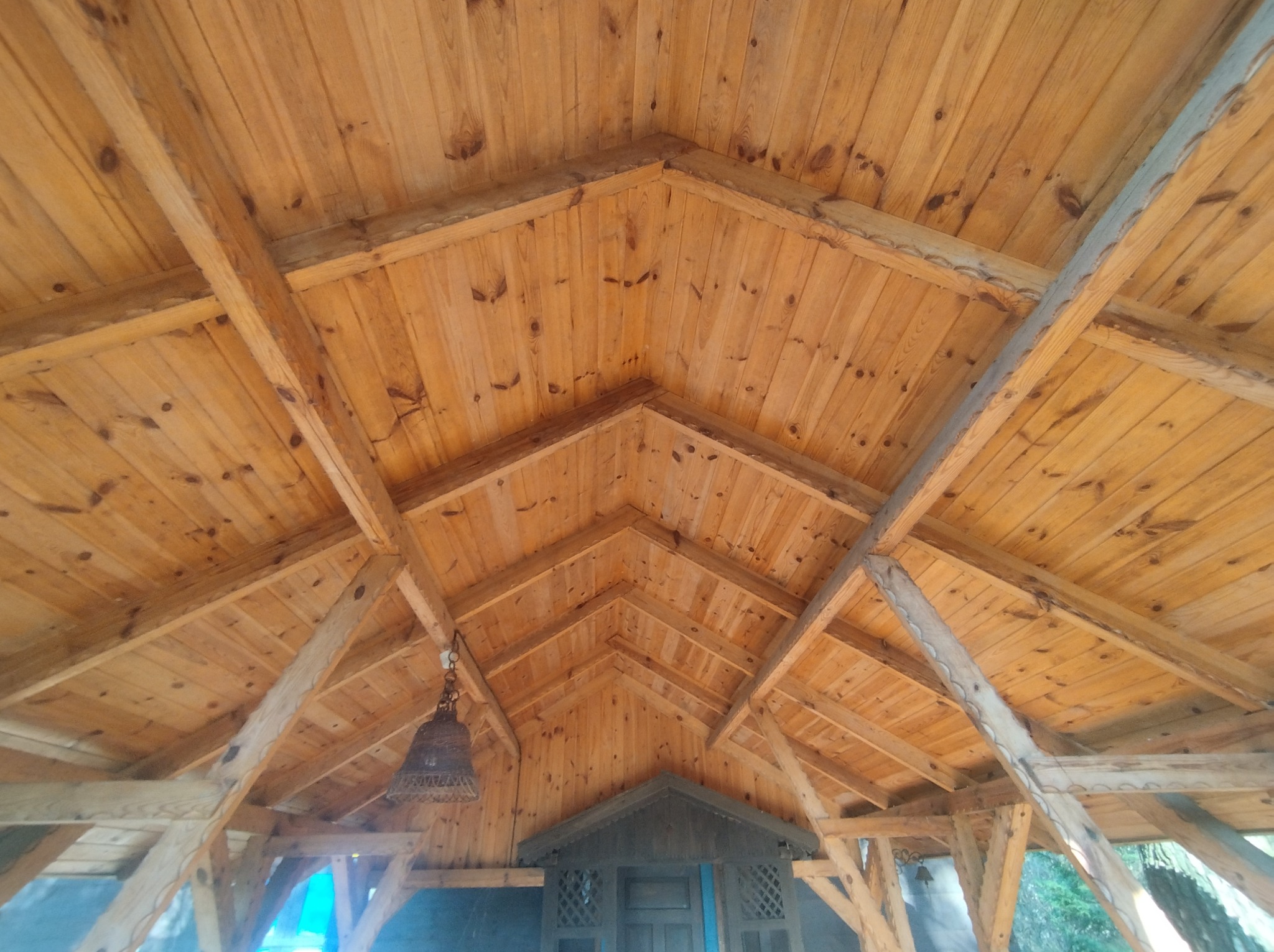 Drewniany sufit z widocznymi belkami konstrukcyjnymi, oświetlony wiszącą lampą w stylu rustykalnym, z fragmentem drzwi w tle.