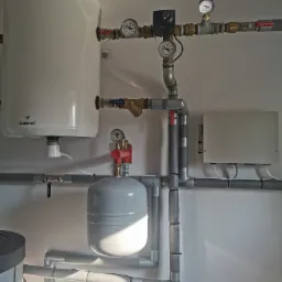 Biały zasobnik ciepłej wody użytkowej z osprzętem, manometrami i rurami w szarej izolacji, zamontowany na białej ścianie w kotłowni.