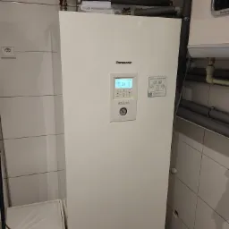 Biała pompa ciepła Panasonic Aquarea w pomieszczeniu z białymi płytkami, widoczne rury instalacyjne i zasobnik ciepłej wody Ariston, na wyświetlaczu pompy widoczna temperatura 31 stopni Celsjusza.