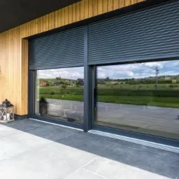 Eleganckie, przesuwne okno z szarymi roletami zewnętrznymi na drewnianej elewacji domu. Widok z zewnątrz na taras z roślinami w donicach.