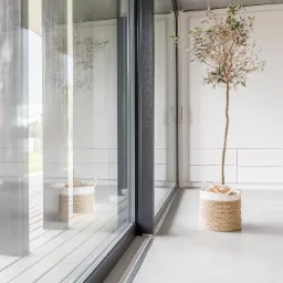 Duże przesuwne okno z czarną ramą, widok na taras i ogr&oacute;d. Minimalistyczne wnętrze z betonowym sufitem i rośliną w plecionym koszu.