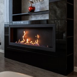 Salon MAKO - Okna Drzwi Bramy i Kominki - Sprzedaż i Montaż - Błyszczący, czarny kominek bio w nowoczesnym salonie z szarą ścianą i drewnianą podłogą. Na kominku dekoracja w kształcie czerwonego jabłka.