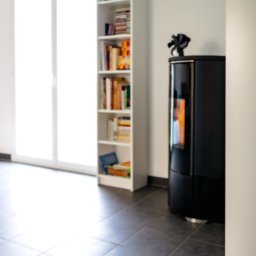 Salon MAKO - Okna Drzwi Bramy i Kominki - Sprzedaż i Montaż - Czarny, nowoczesny kominek wolnostojący w jasnym wnętrzu z półką na książki i siedziskiem przy oknie. Minimalistyczny design, szare płytki na podłodze.