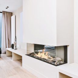 Salon MAKO - Okna Drzwi Bramy i Kominki - Sprzedaż i Montaż - Nowoczesny kominek narożny z paleniskiem osłoniętym szkłem, wkomponowany w minimalistyczne, białe wnętrze salonu z drewnianą podłogą i subtelnymi dekoracjami.