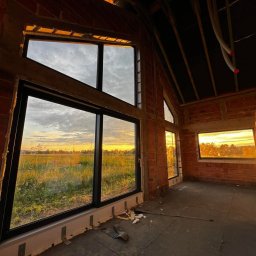 Salon MAKO - Okna Drzwi Bramy i Kominki - Sprzedaż i Montaż - Surowy stan wnętrza z dużymi oknami z czarnymi ramami, przez które widać zachodzące słońce nad polem. Widoczne ceglane ściany i elementy instalacji.