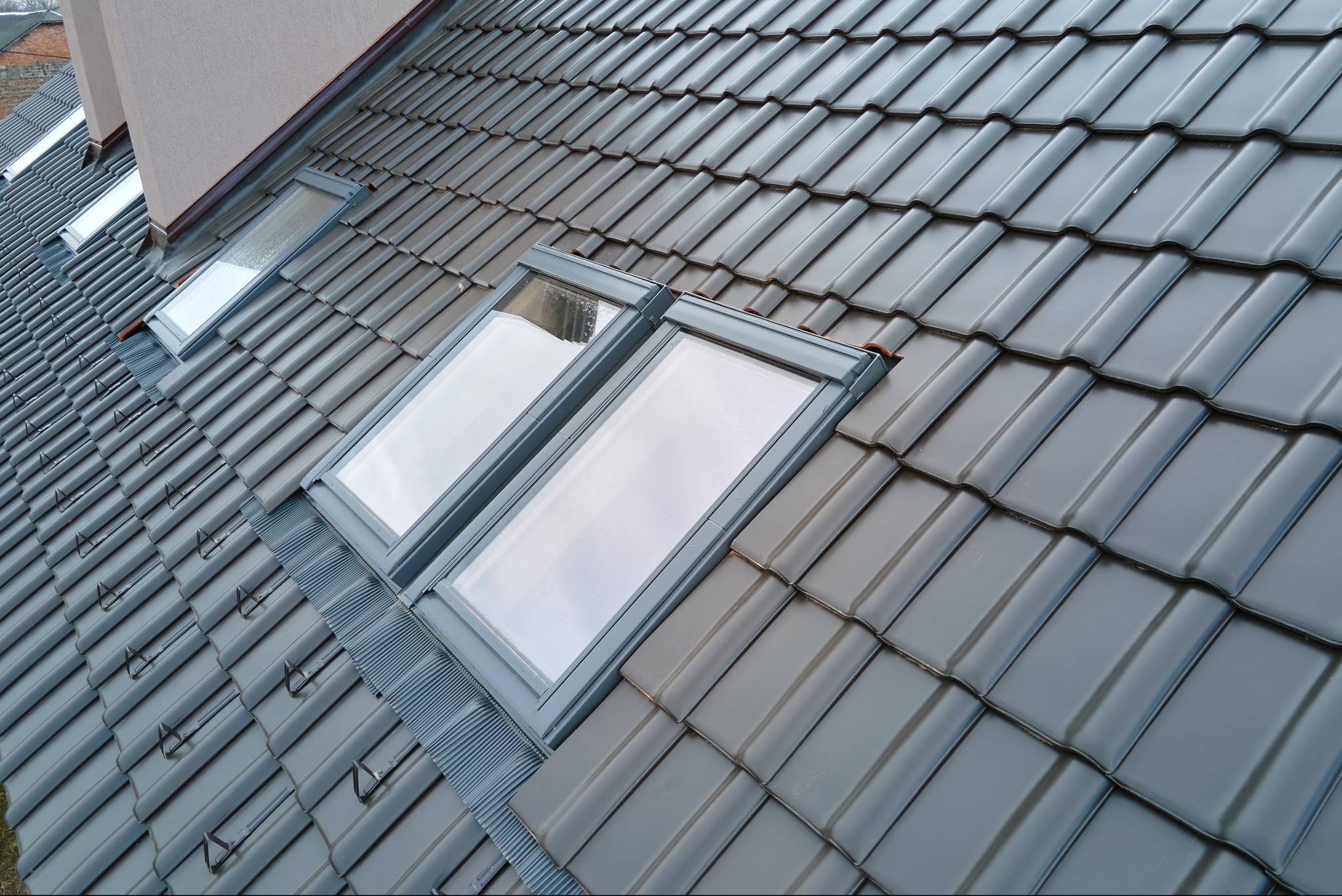Dwa okna dachowe w szarej ramie, zamontowane na ciemnoszarym dachu z dachówki ceramicznej. Widoczny fragment komina w tle.