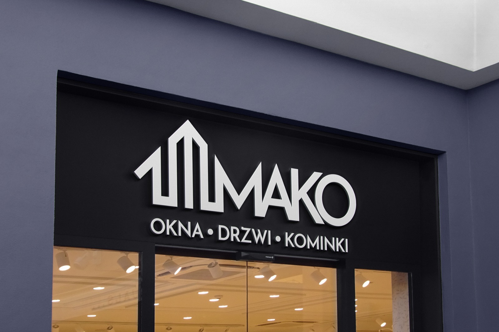 Elegancki szyld firmy MAKO nad wejściem, oferującej okna, drzwi i kominki. Białe litery na czarnym tle, minimalistyczny design, widoczne oświetlenie wnętrza sklepu.