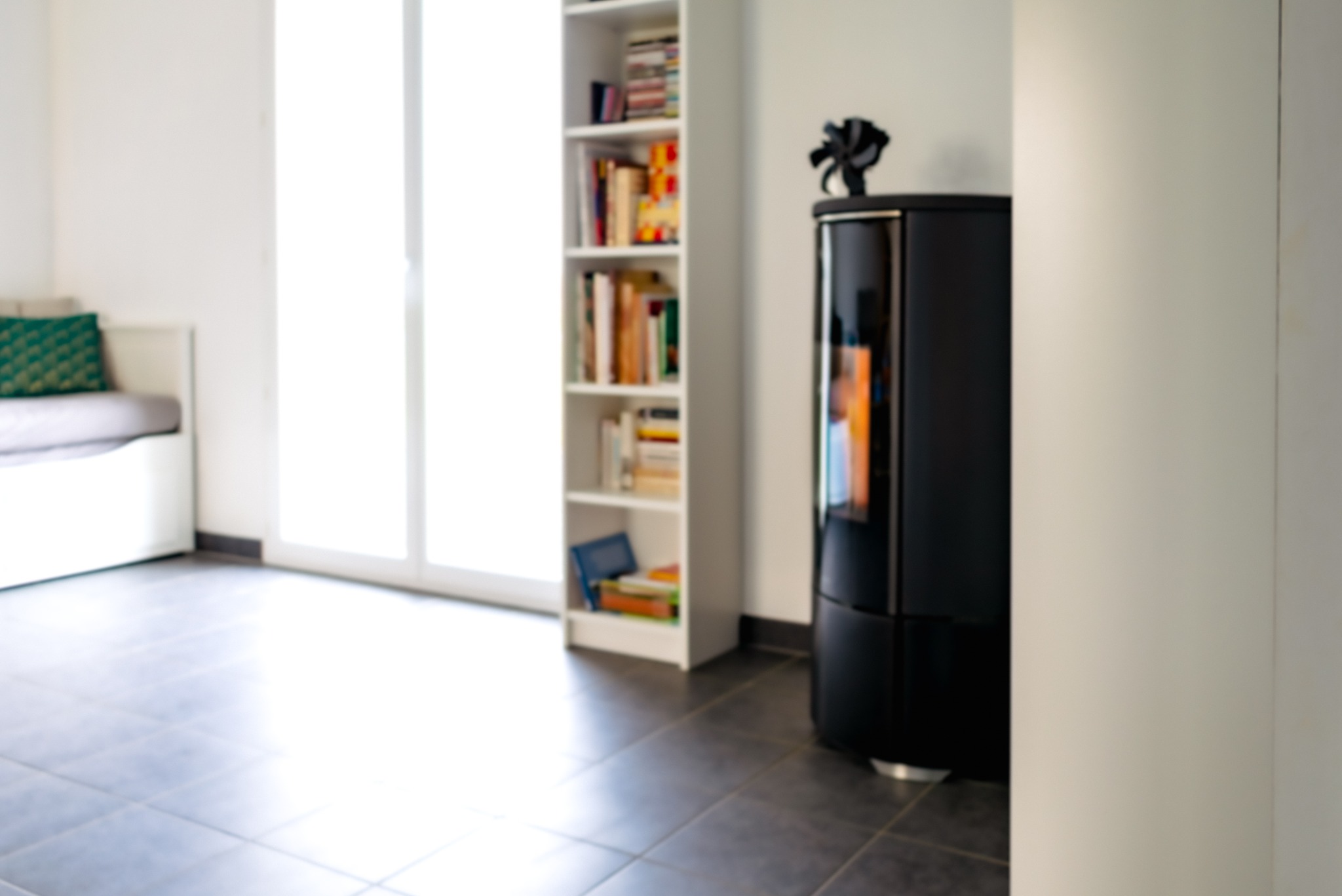 Czarny, nowoczesny kominek wolnostojący w jasnym wnętrzu z półką na książki i siedziskiem przy oknie. Minimalistyczny design, szare płytki na podłodze.