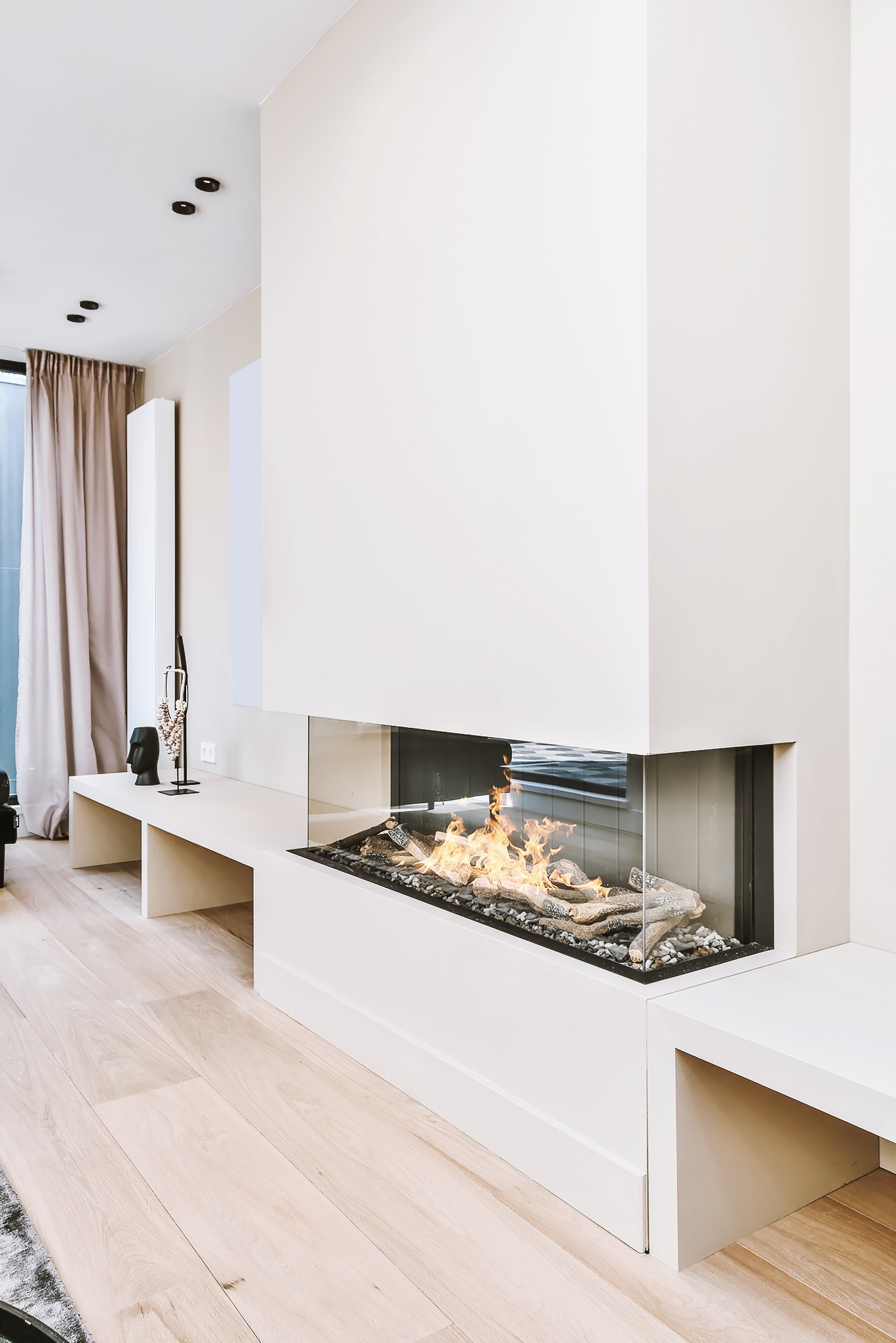 Nowoczesny kominek narożny z paleniskiem osłoniętym szkłem, wkomponowany w minimalistyczne, białe wnętrze salonu z drewnianą podłogą i subtelnymi dekoracjami.