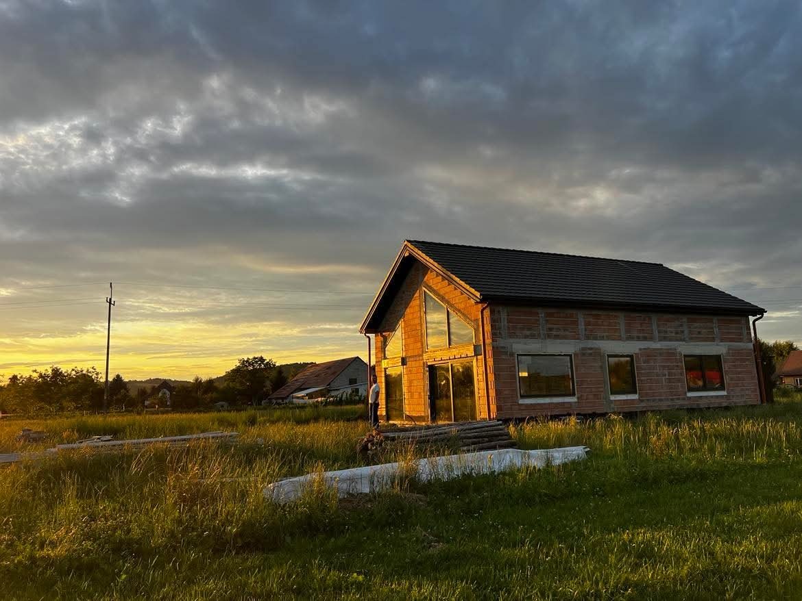 Surowy stan domu jednorodzinnego z dużymi oknami w Zakliczynie, w tle zachodzące słońce i pochmurne niebo. Widoczne cegły i konstrukcja dachu.