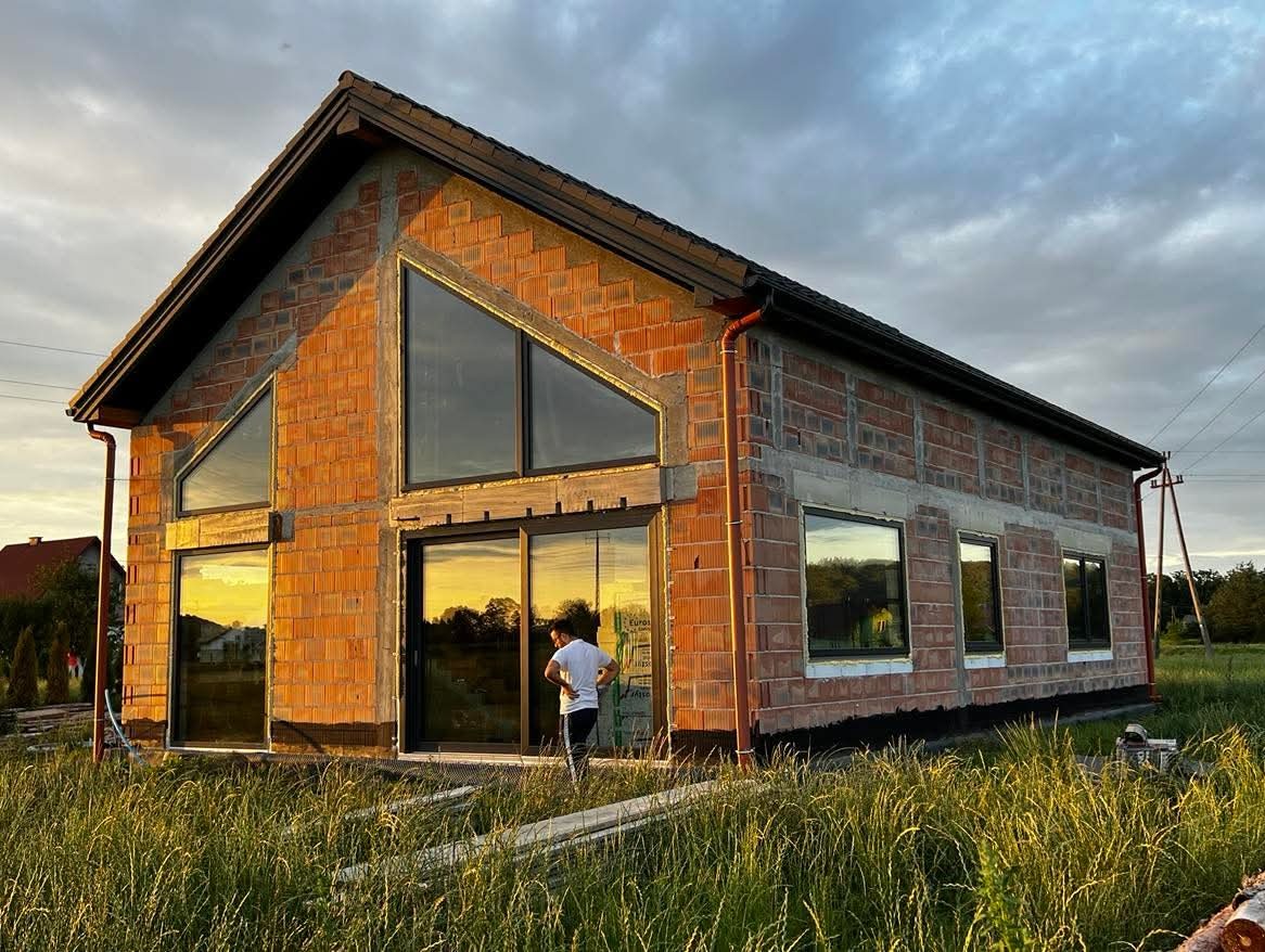 Surowy budynek z cegły z dużymi oknami i przesuwnymi drzwiami. Widoczne odblaski zachodzącego słońca w szybach, wysoka trawa, osoba w pobliżu budynku.