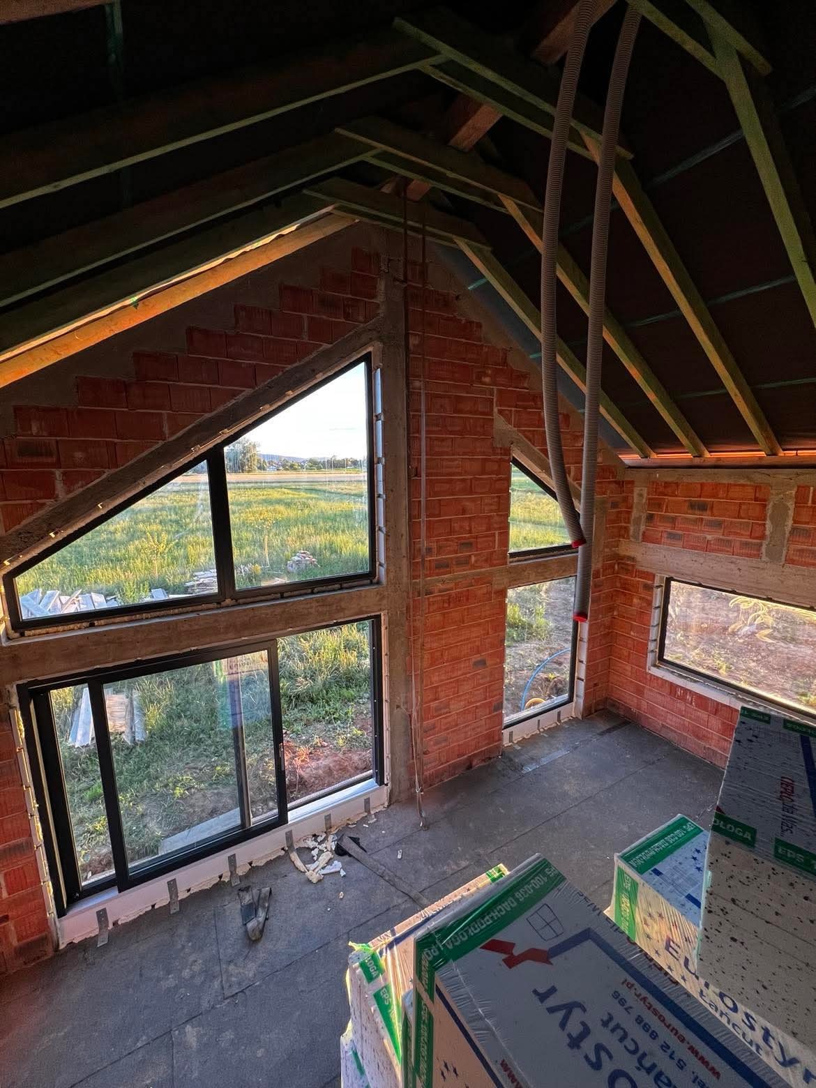 Surowy stan wnętrza z oknami o nietypowych kształtach w murowanym budynku. Widoczne materiały budowlane i elementy konstrukcji dachu. Krajobraz za oknem.