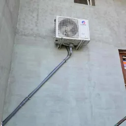 Zewnętrzna jednostka klimatyzacji marki Gree zamontowana na szarej, chropowatej ścianie budynku z widocznym odprowadzeniem skroplin w postaci szarej rury PCV, zabezpieczonej obejmami.