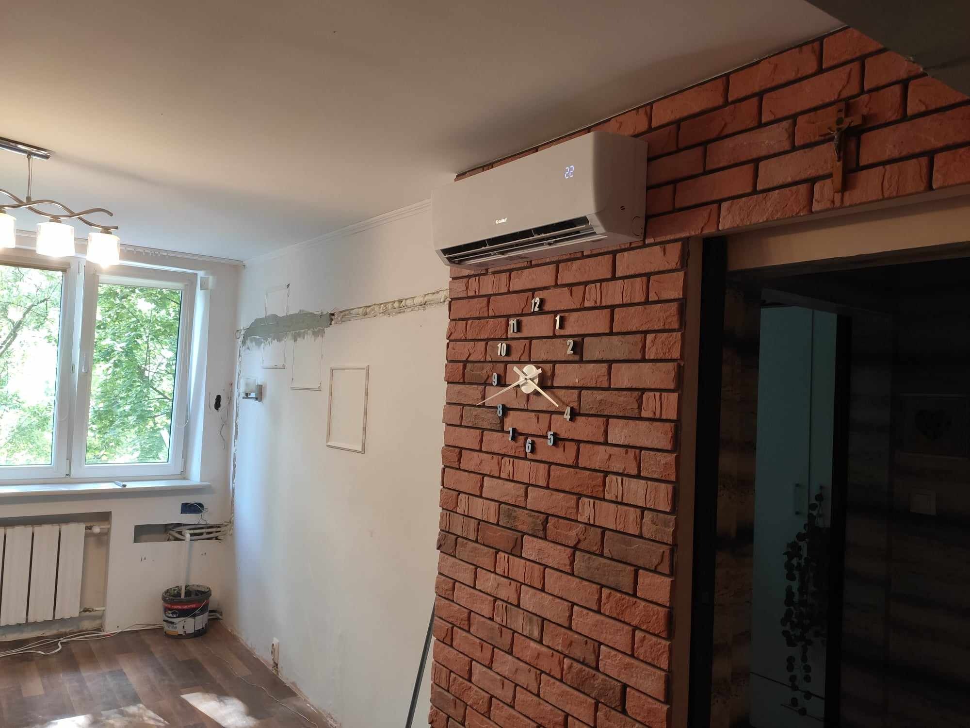 Biały klimatyzator zamontowany na ścianie z czerwonej cegły w pokoju z oknem, widoczne niedokończone prace wykończeniowe i wiadro farby.