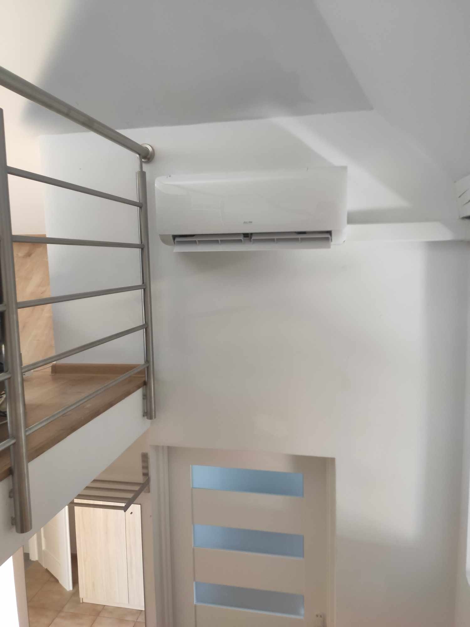 Biały klimatyzator na ścianie w jasnym wnętrzu, widok z góry na drzwi z przeszkleniami i metalową balustradę schodów.