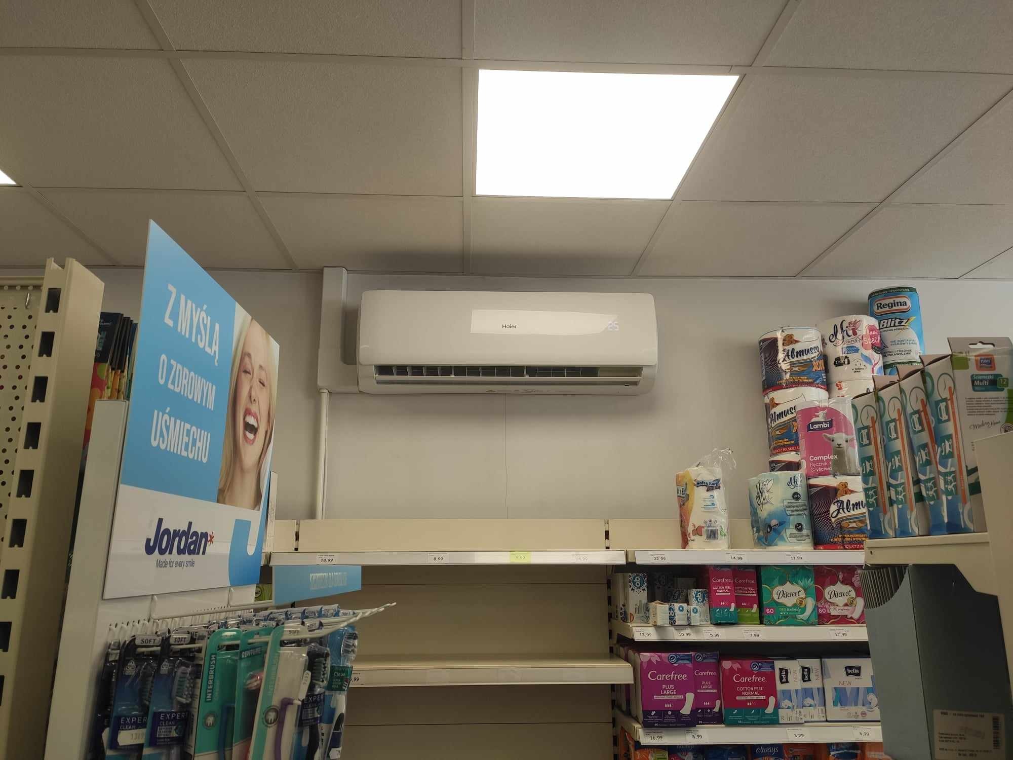 Biały klimatyzator Haier zamontowany w sklepie, widoczny na tle półek z towarem, oświetlenie sufitowe.
