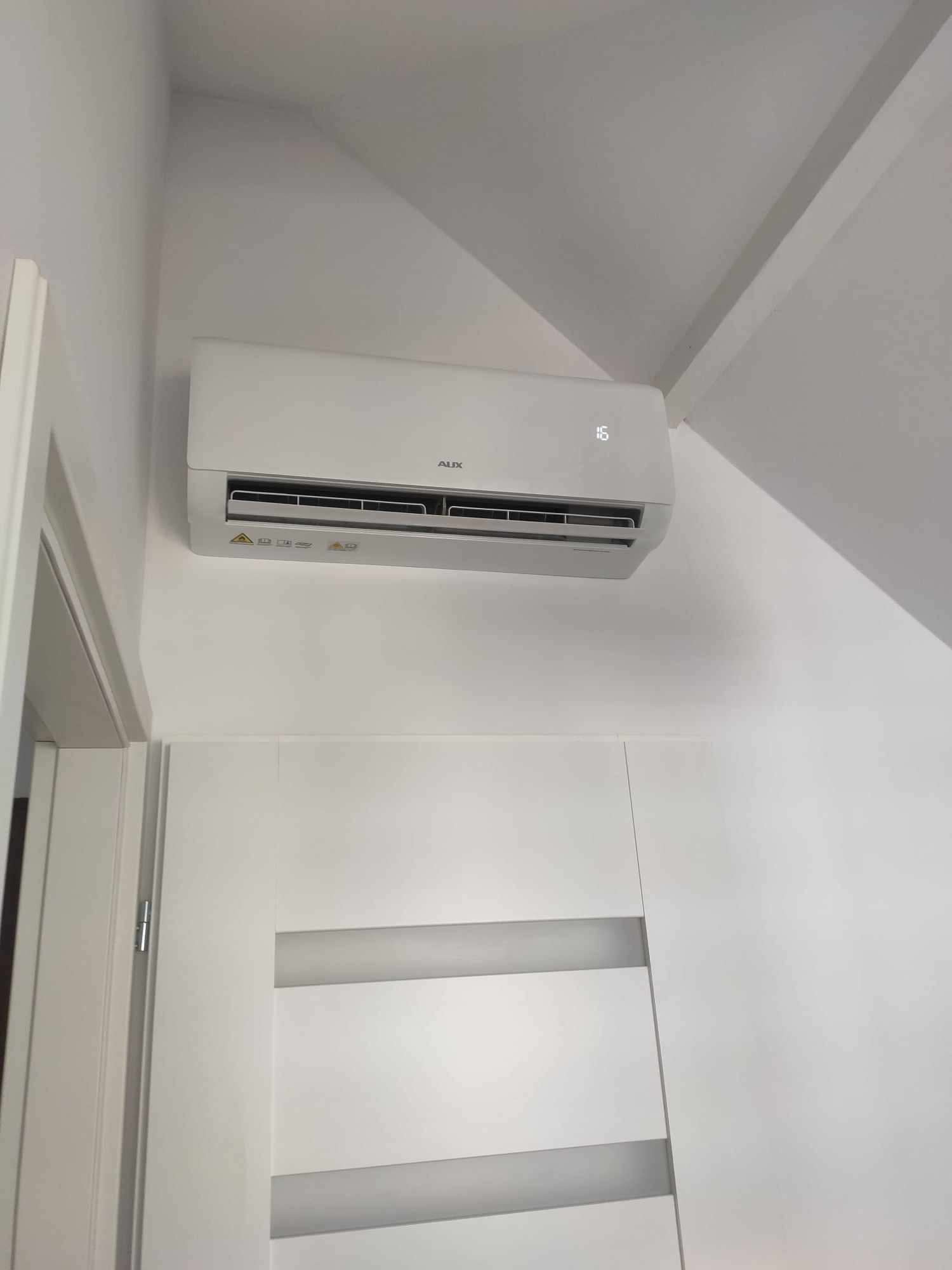 Biały klimatyzator AUX zamontowany na ścianie nad białą szafą w pomieszczeniu o białych ścianach i suficie.
