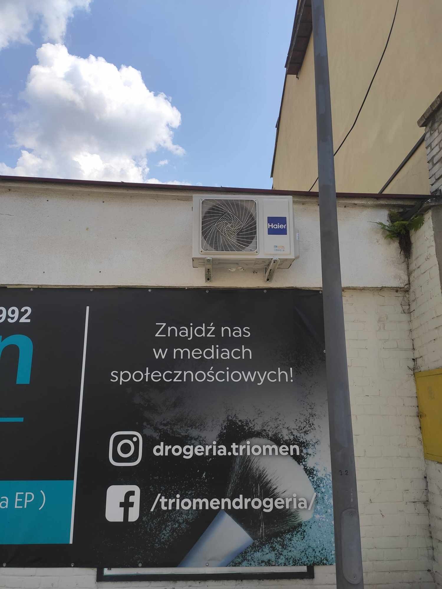 Zamontowana jednostka zewnętrzna klimatyzacji Haier na ścianie budynku z reklamą drogerii, widok z dołu w słoneczny dzień.