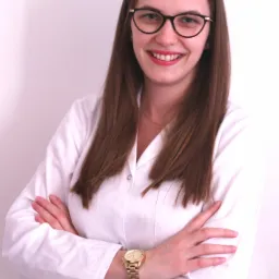 Uśmiechnięta kobieta w okularach i białym uniformie medycznym, ze złotym zegarkiem na ręce, pozuje z założonymi rękami na białym tle.