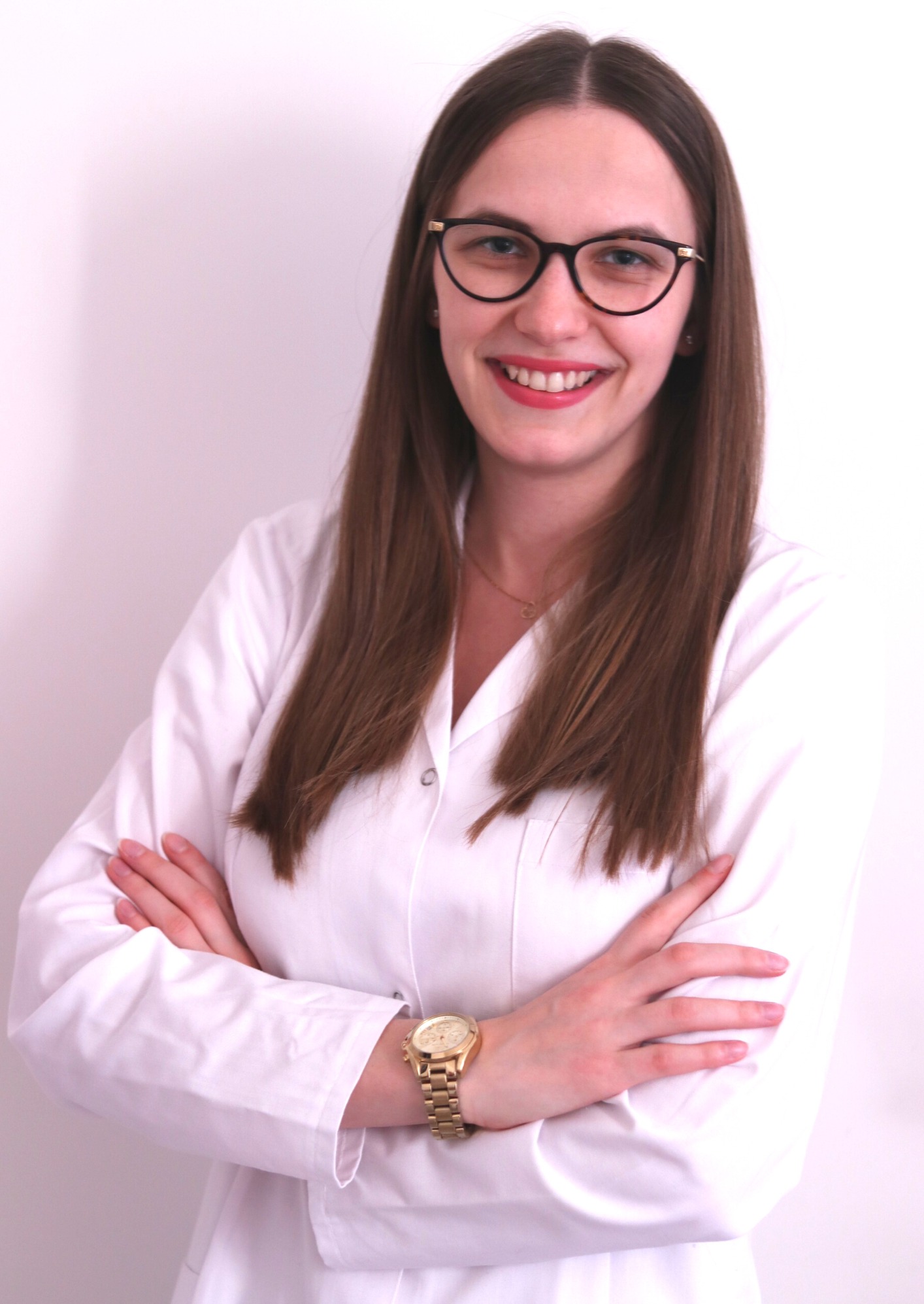 Uśmiechnięta kobieta w okularach i białym uniformie medycznym, ze złotym zegarkiem na ręce, pozuje z założonymi rękami na białym tle.