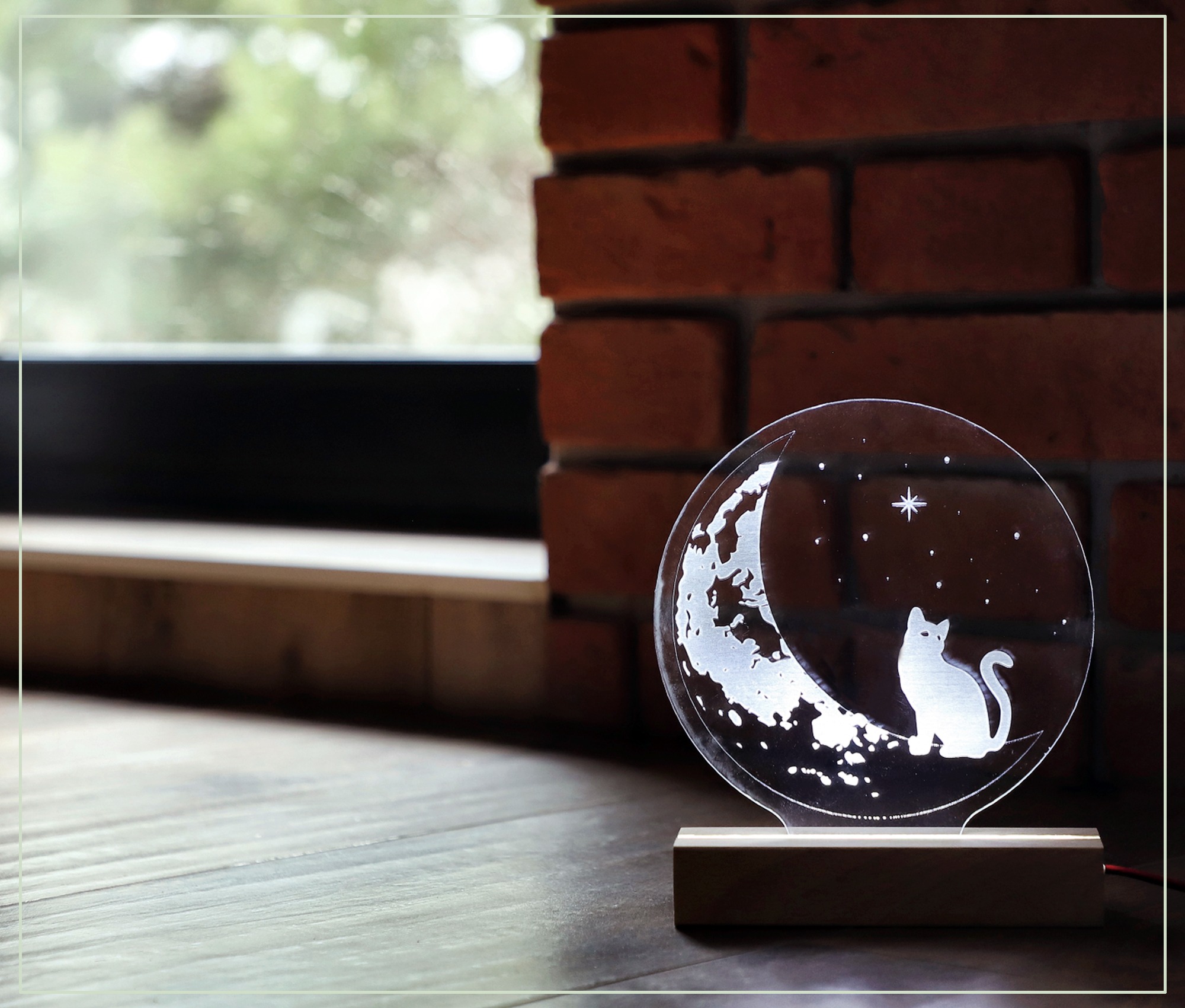 Podświetlana, okrągła lampka nocna z wygrawerowanym motywem kota siedzącego na księżycu i gwiazd, stojąca na drewnianej podstawie, na tle ceglanej ściany i okna.
