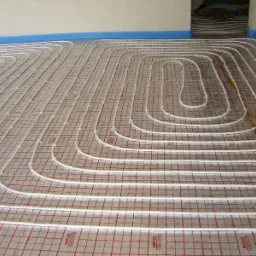 Białe rury grzewcze podłogowe ułożone spiralnie na folii z czerwoną siatką, widoczne mocowania rur do podłoża, przygotowane do zalania wylewką.