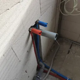 Fragment ściany z betonu komórkowego z przejściami rur instalacji wodnej i kanalizacyjnej w kolorach niebieskim i czerwonym, oraz szara rura kanalizacyjna wystająca ze ściany.