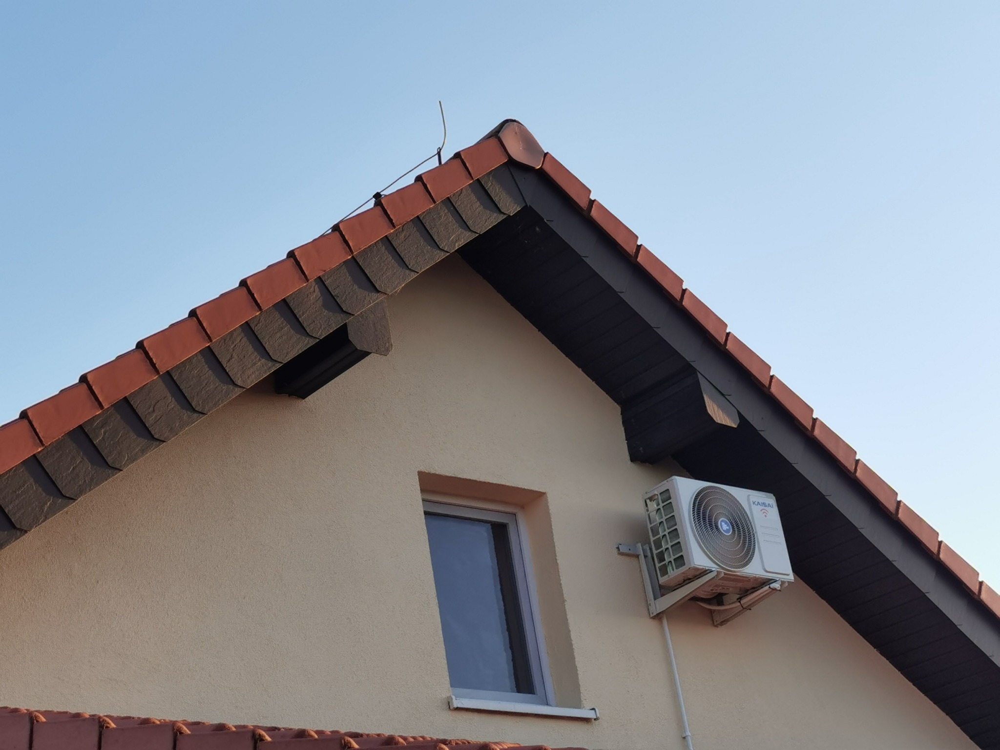Zewnętrzna jednostka klimatyzacji Kaisai zamontowana na ścianie domu z beżową elewacją, widoczna z perspektywy żabiej z czerwoną dachówką i anteną na dachu.
