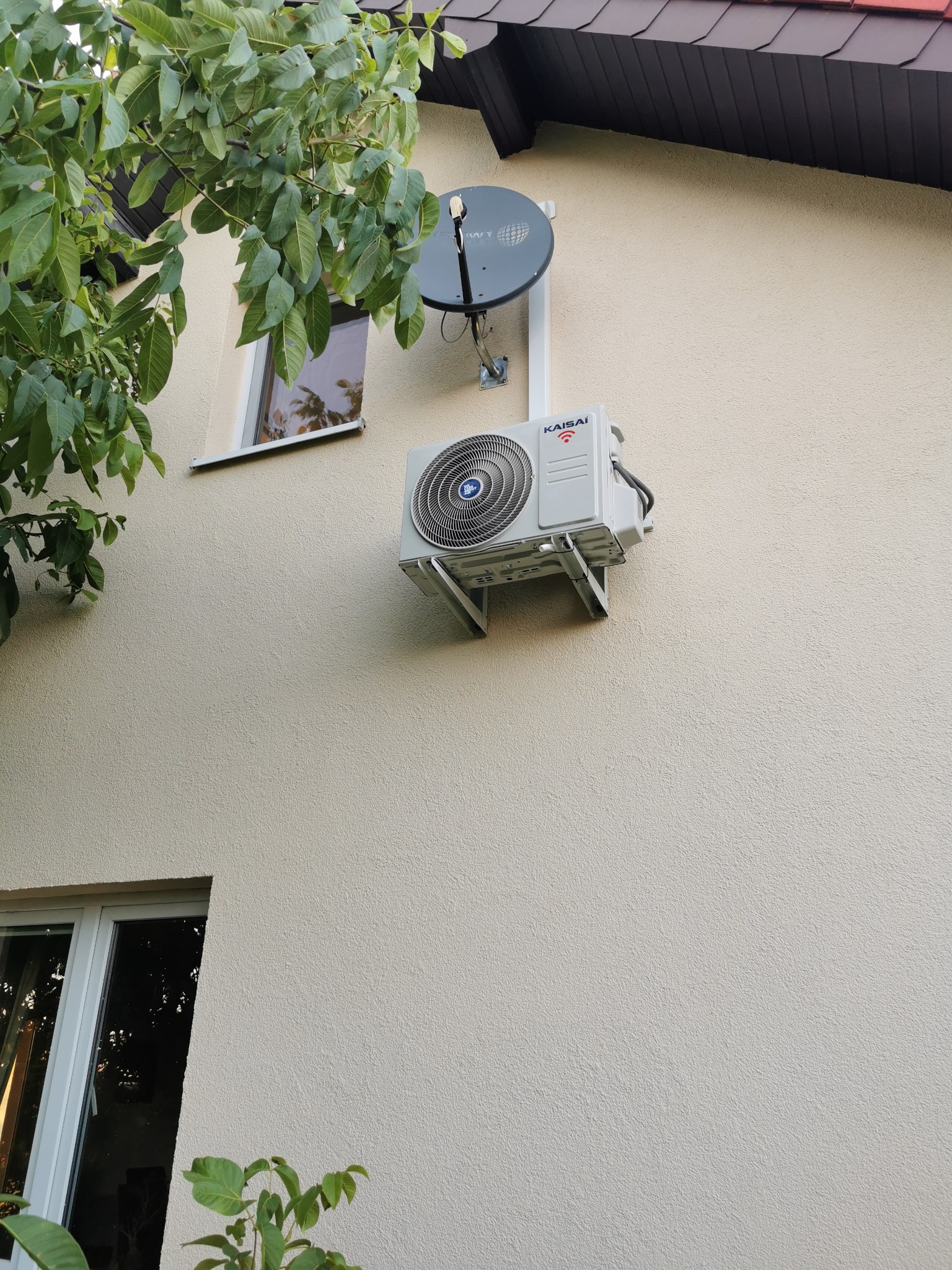 Zewnętrzna jednostka klimatyzatora Kaisai zamontowana na beżowej elewacji domu, widoczna antena satelitarna i fragment drzewa.