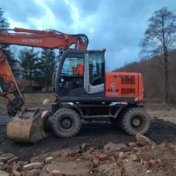 Pomarańczowo-szary kołowy koparka Hitachi Zaxis 140W na nierównym terenie z gruzem i ziemią, widoczny operator w pomarańczowej kamizelce, drzewa w tle.